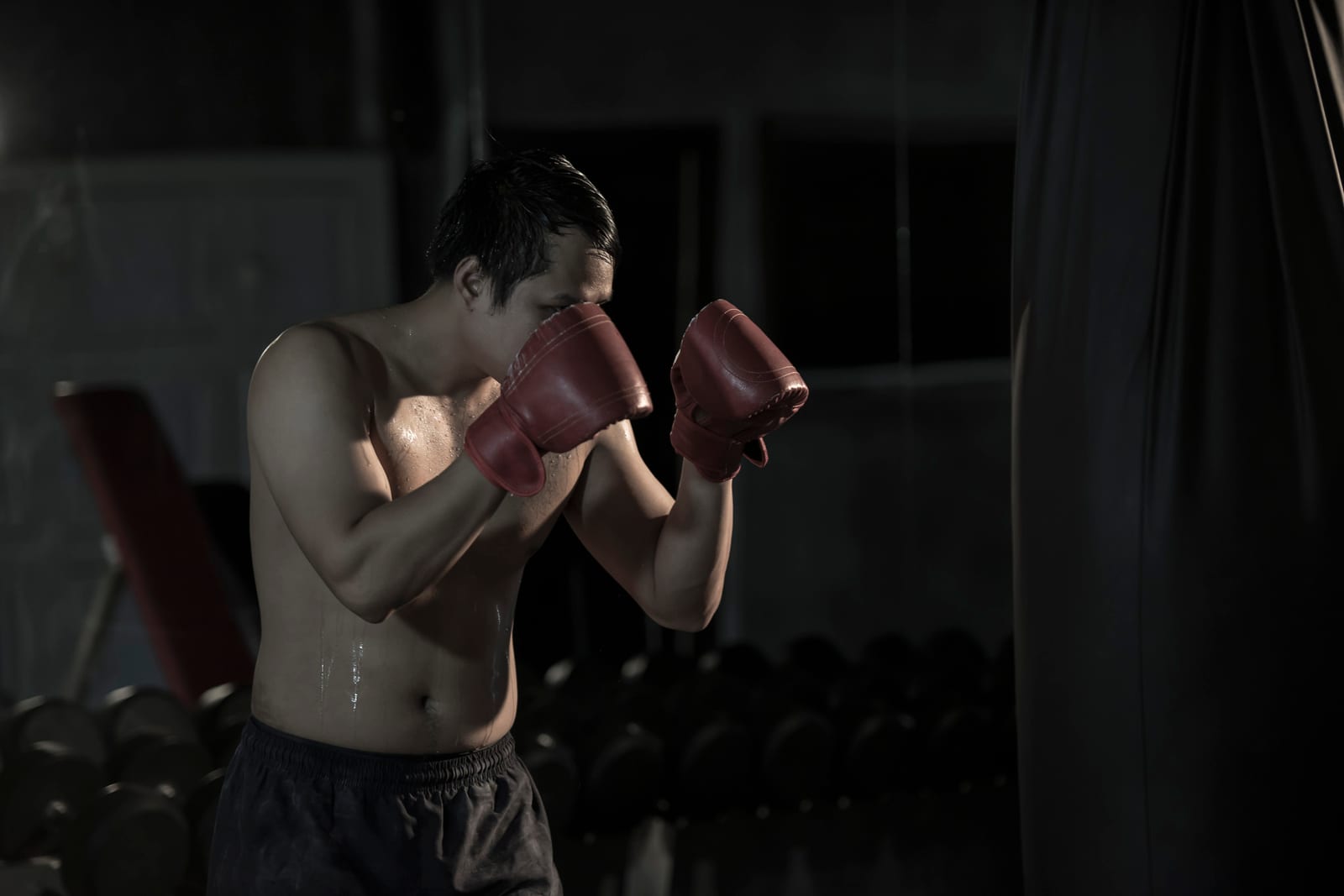 A man in dark red boxing gloves holding a tight guard in a dim gym.