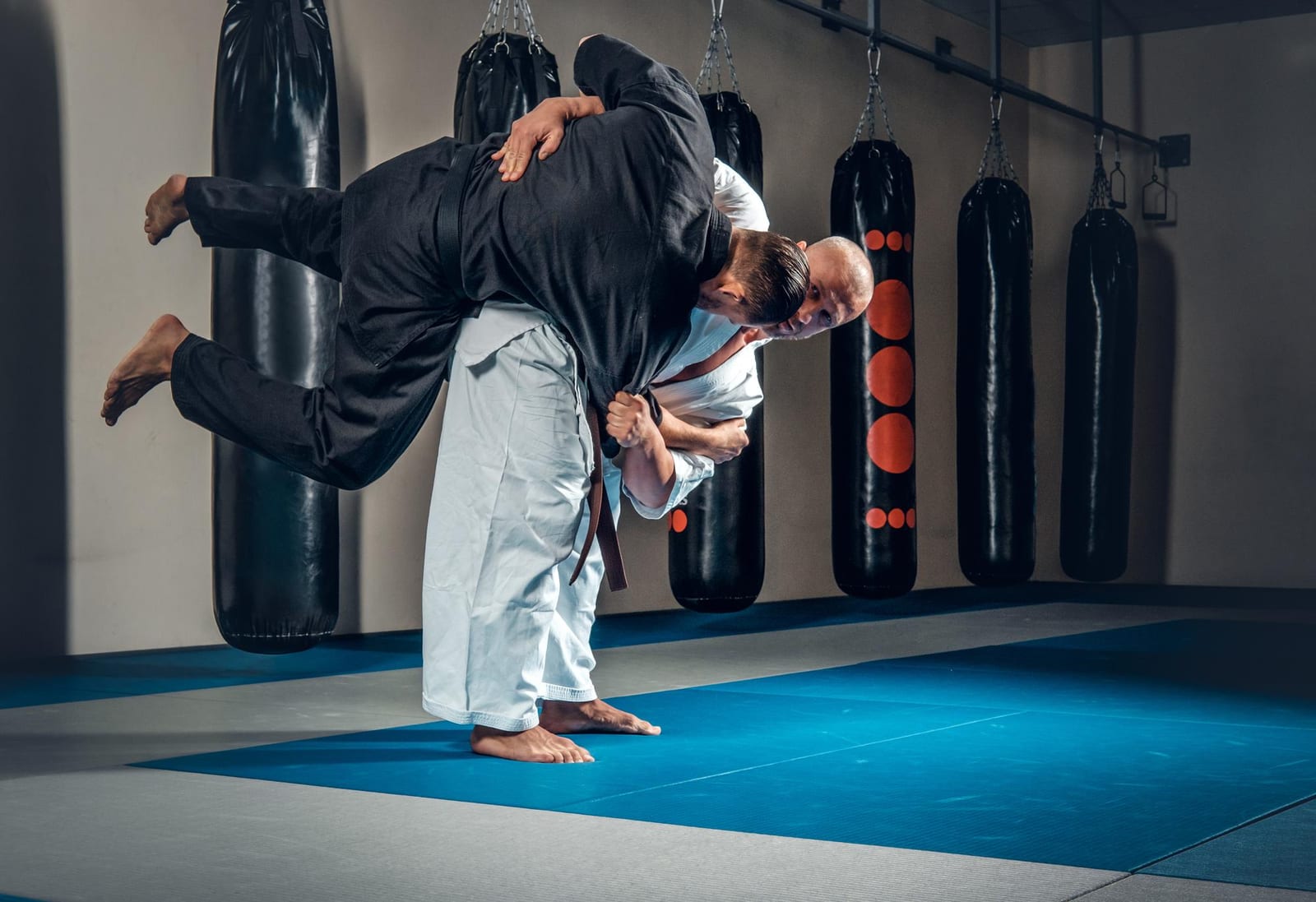 A judoka in a white gi lifting a partner for a throw beside a row of heavy bags.