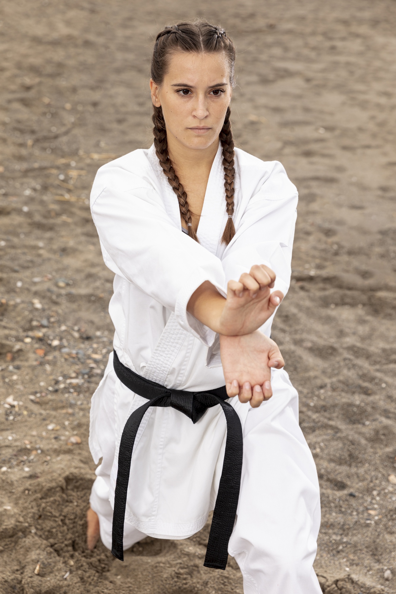 A woman in a white gi and black belt kneeling on sand, holding a low block.