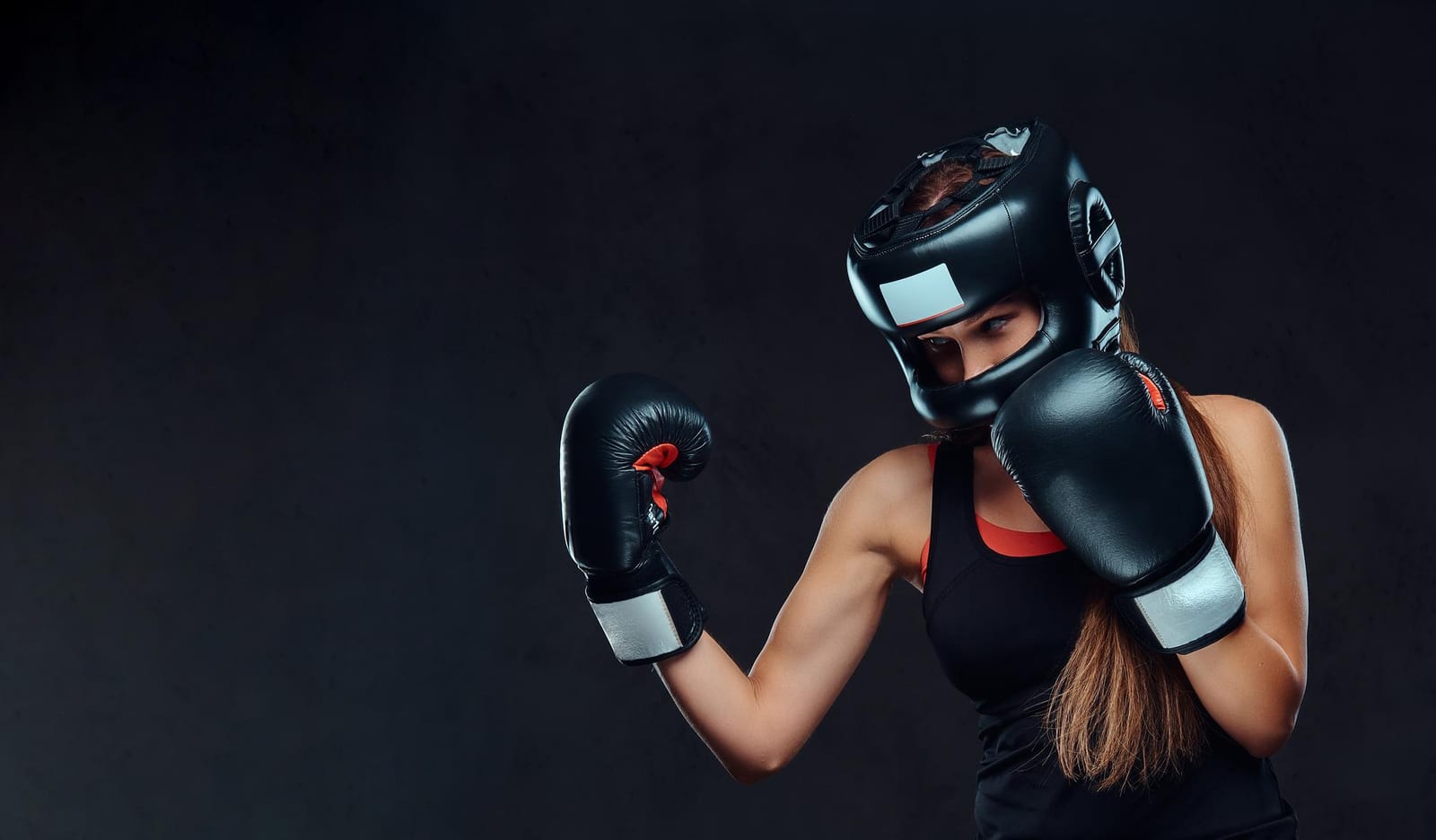 A woman in black headgear and boxing gloves holding her guard against a dark background.