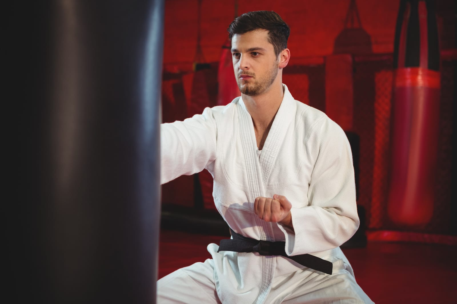 A man in a white gi and black belt punching from a stance in a red-lit gym full of bags.